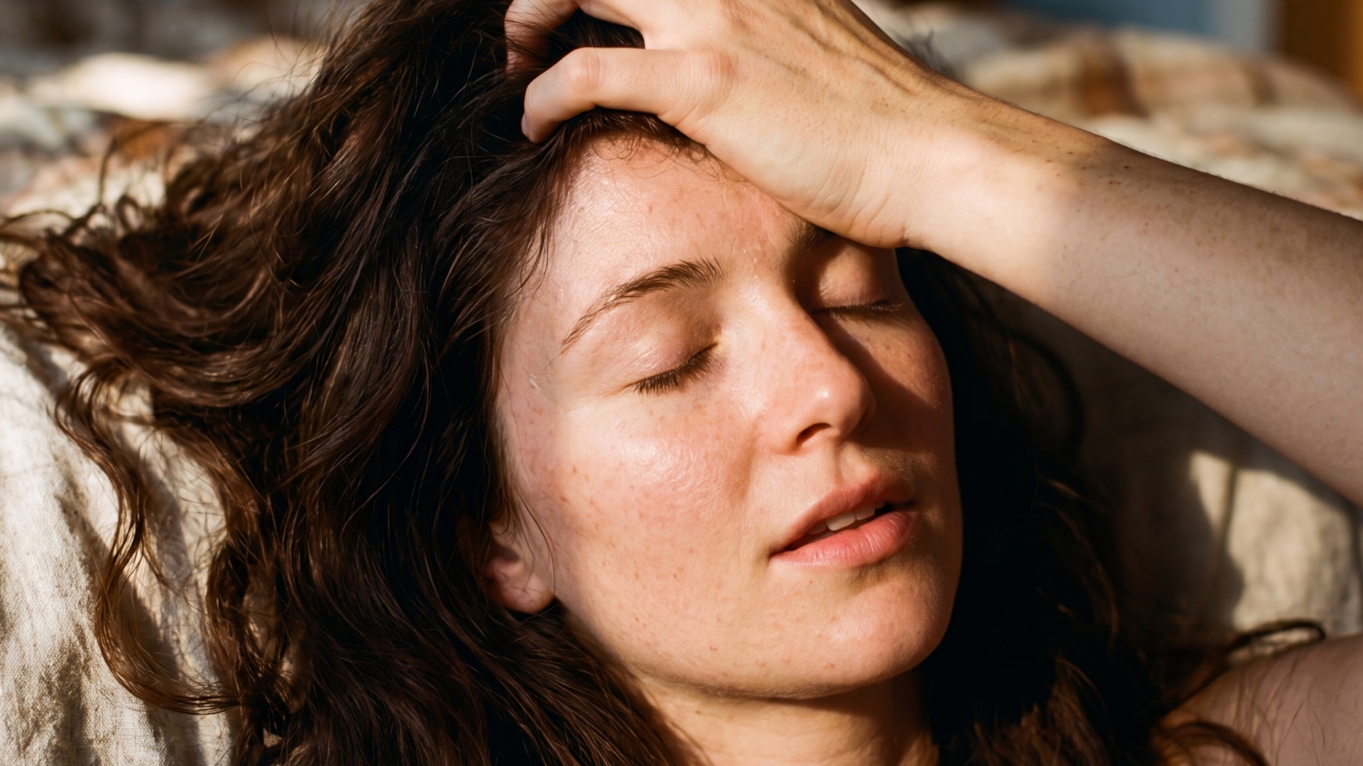 A white brunette woman with freckles lies back in bed, her eyes closed and her hand in her hair