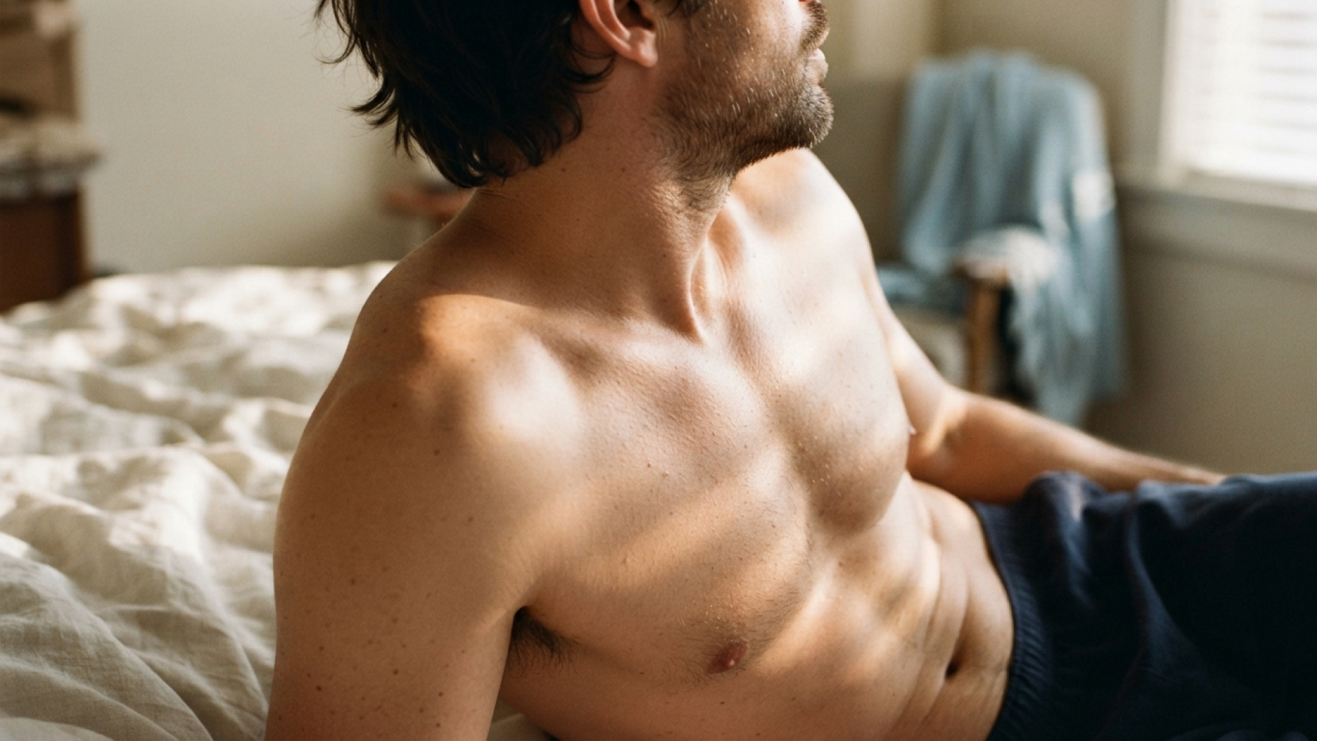 A white man with dark hair lies topless in bed. He has navy blue jogging bottoms on and his cream bedding is crumpled.