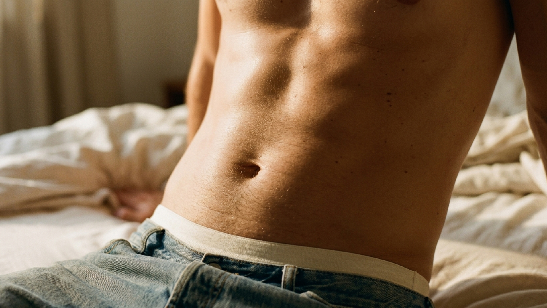Close-up of a man's torso as he sits on a bed. He's wearing jeans.