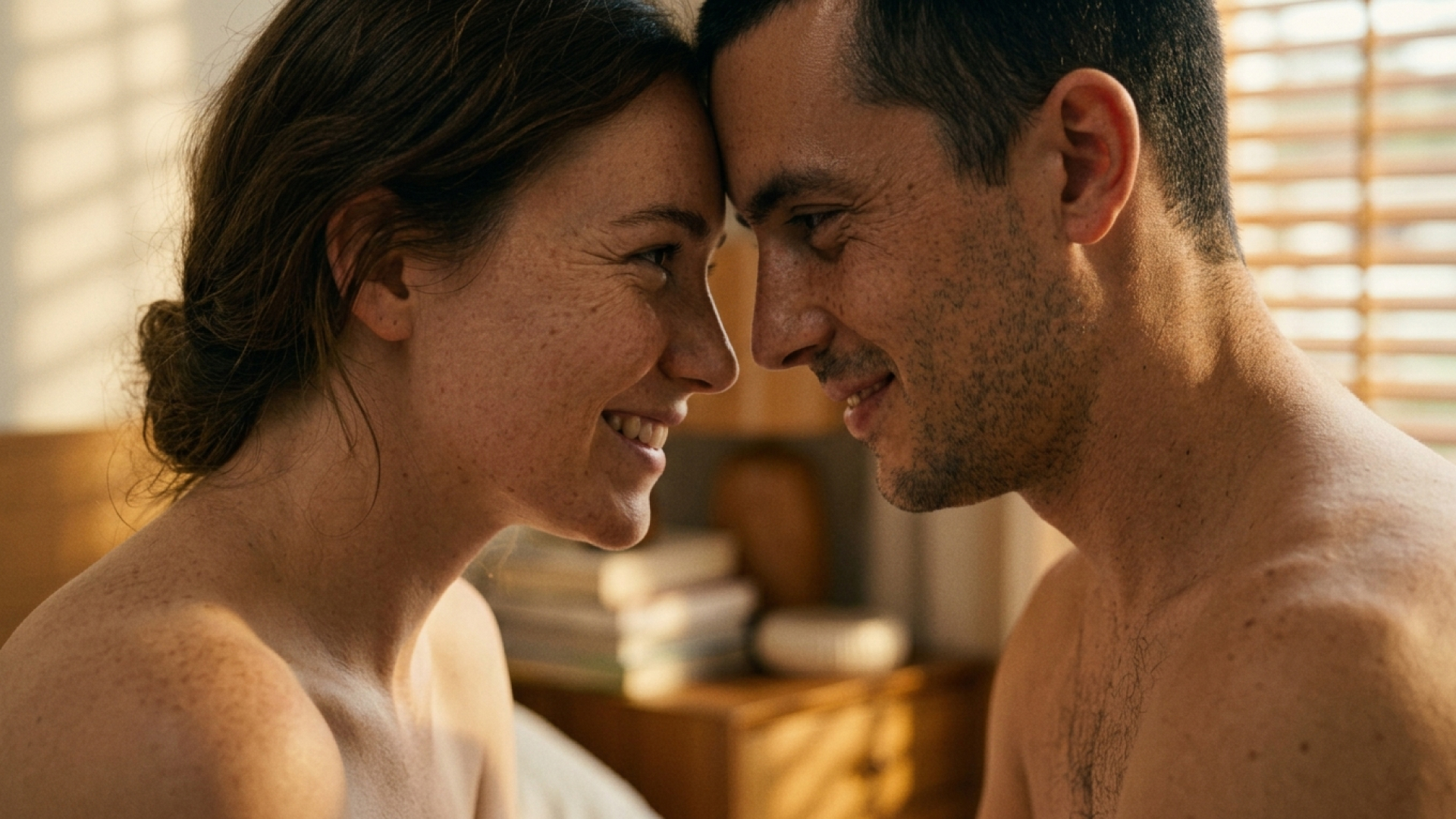 A white couple in their 30s or 40s face each other, smiling. They appear to be naked, shown from the shoulders up, and are touching foreheads. They both have brown hair.