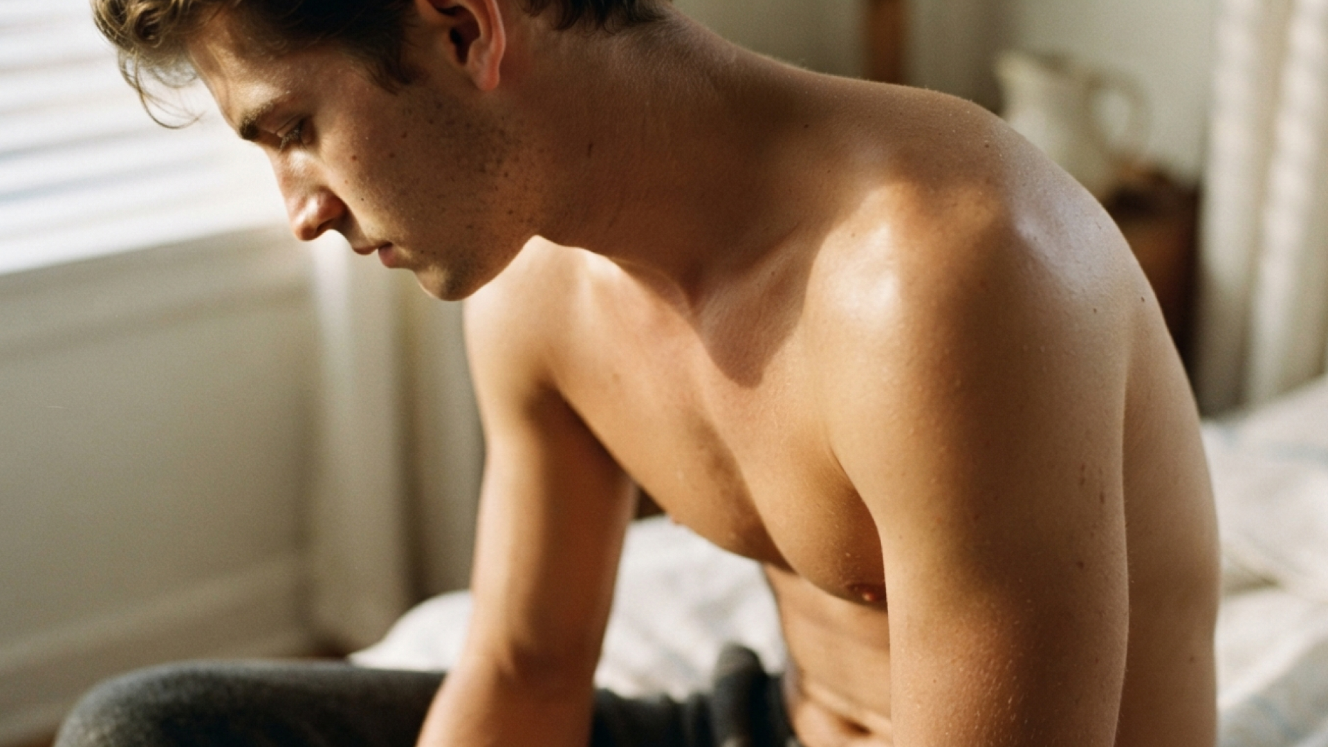 A topless white man in his 20s sits on a bed looking pensively at the floor