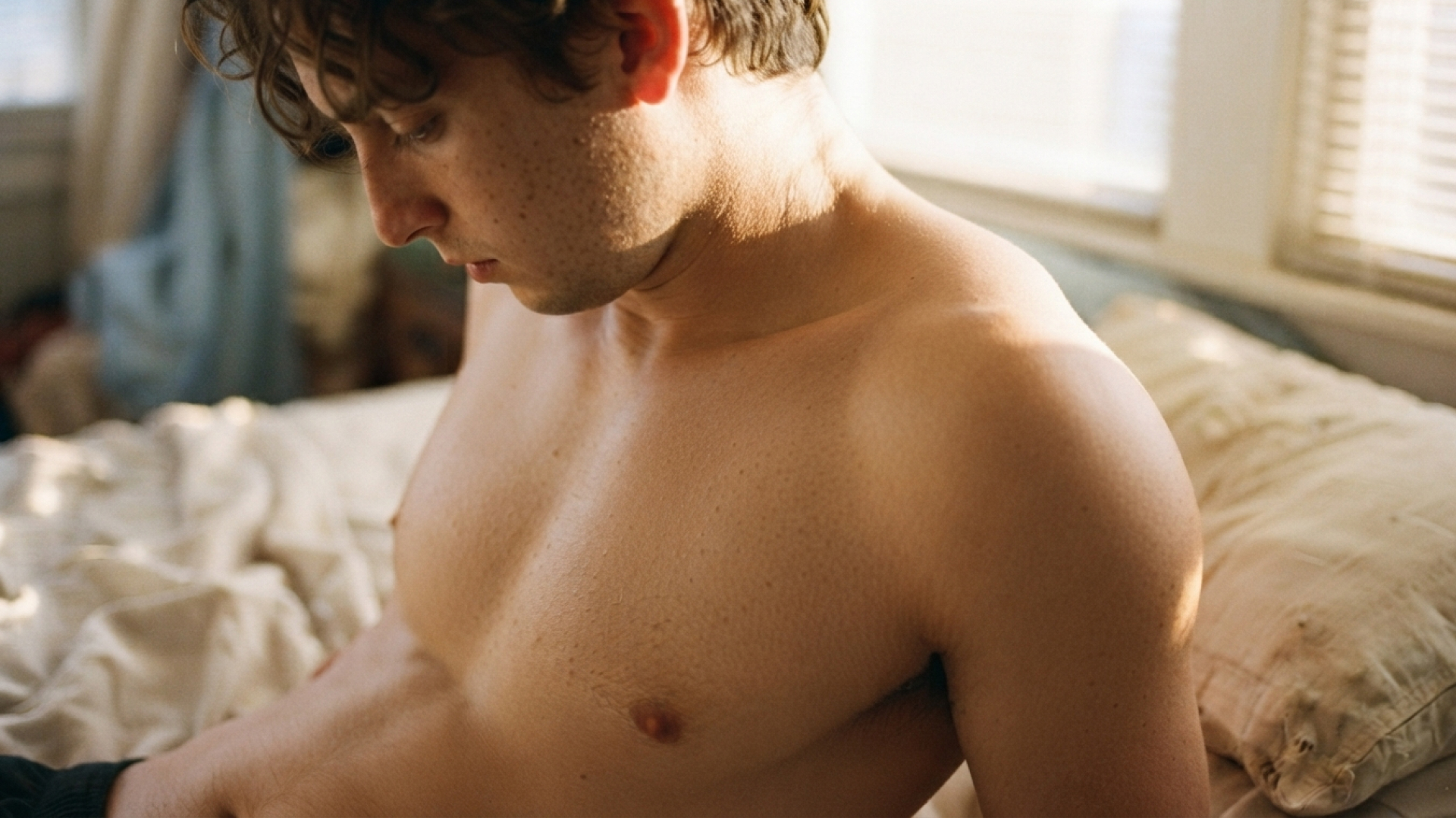 A topless white man with longish hair sits up in bed, looking sadly down