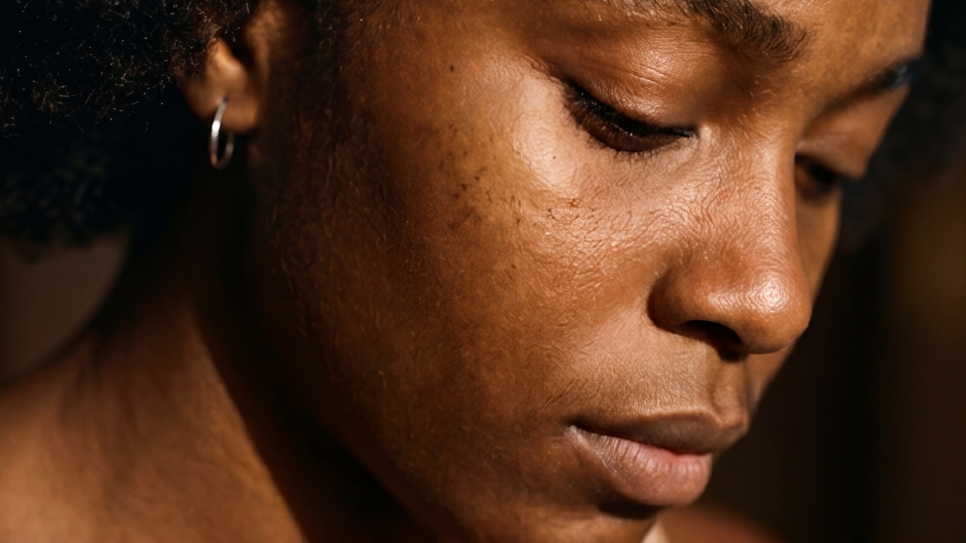 A Black woman with light freckles and silver hoop earrings looks pensively down