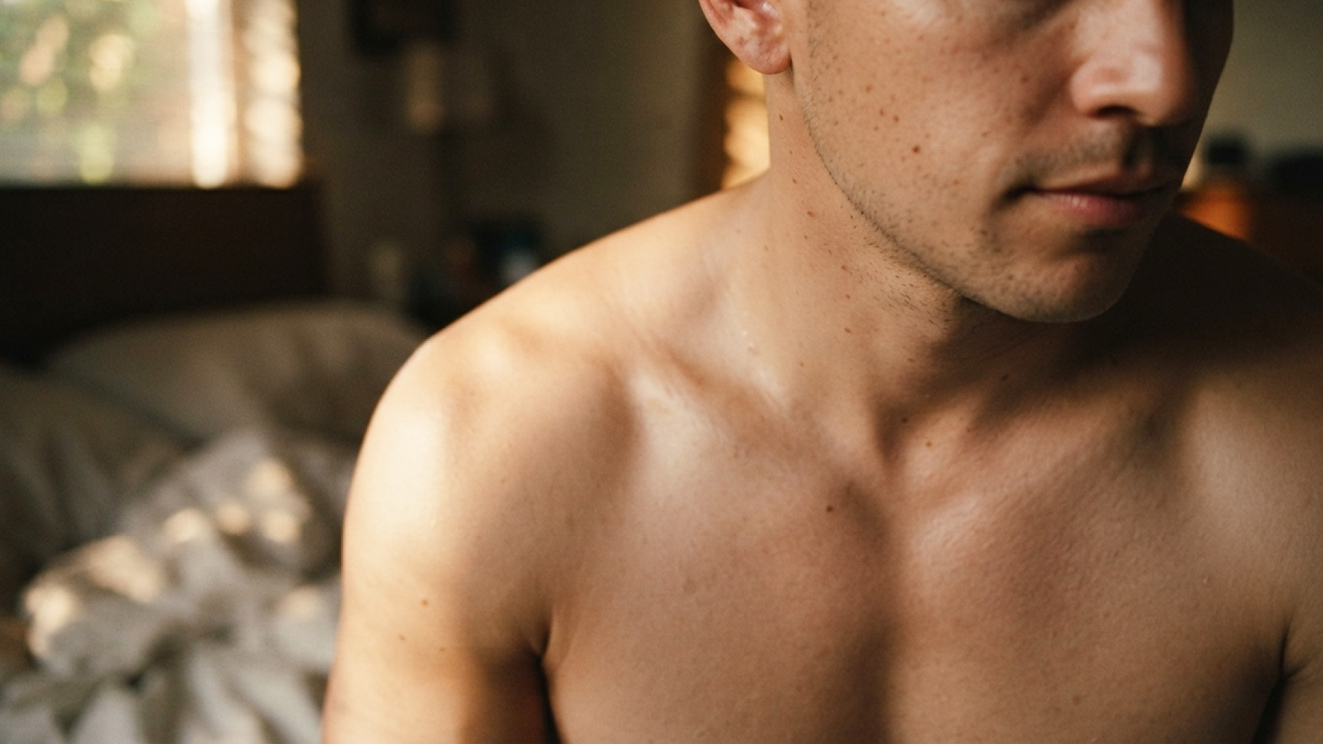 A white man with light stubble is sat at the end of his bed; the image is cropped from his chest to his nose and he appears to be naked