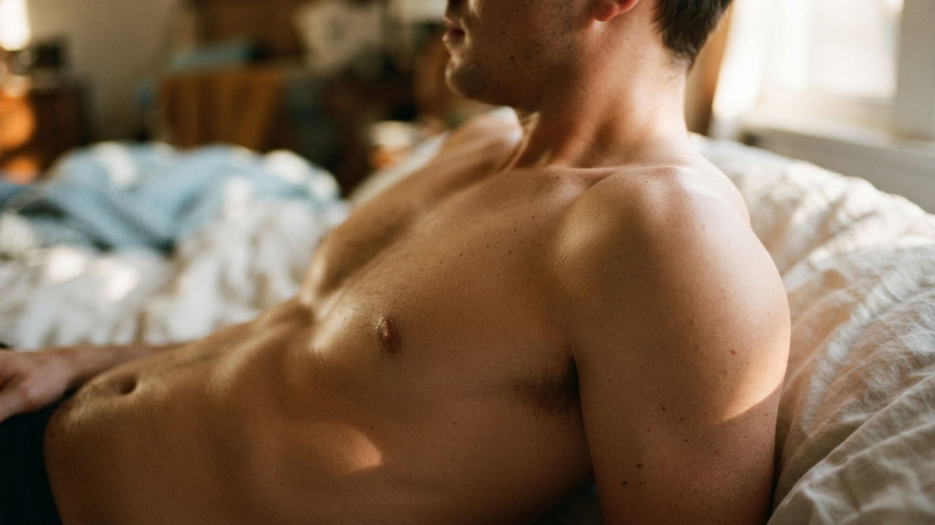 A tanned man is topless in bed, leaning up on his elbows. The background is blurred.