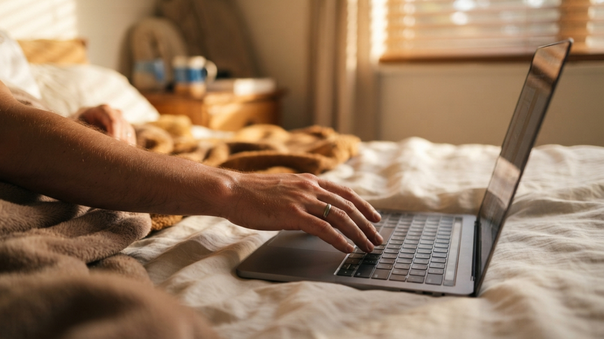 A white man's arm reaches to type on a laptop, which rests on a bed