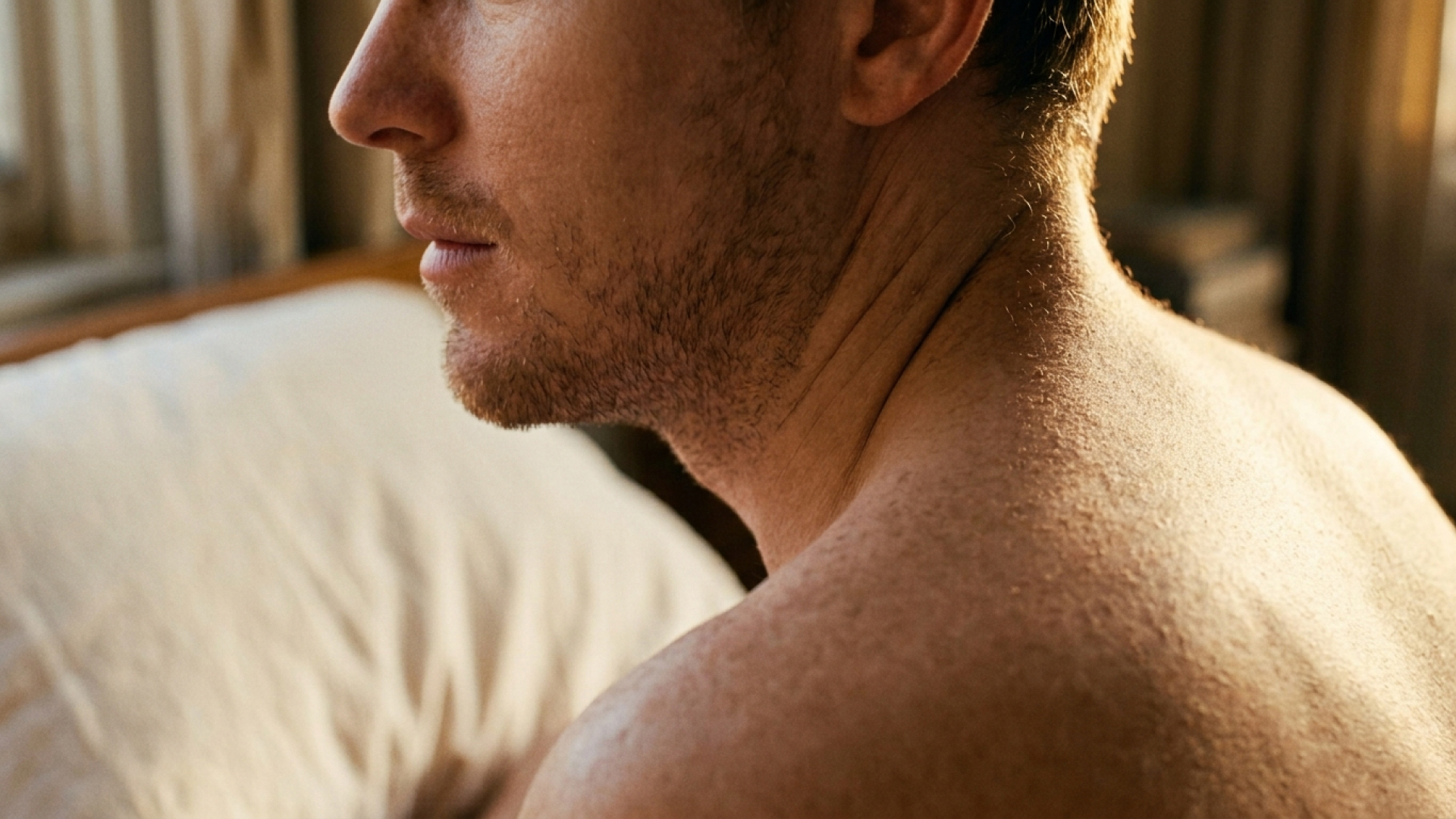 Close-up of a white man with stubble, shown from shoulders to nose. He's looking to the side and you can see his pillow in the background.