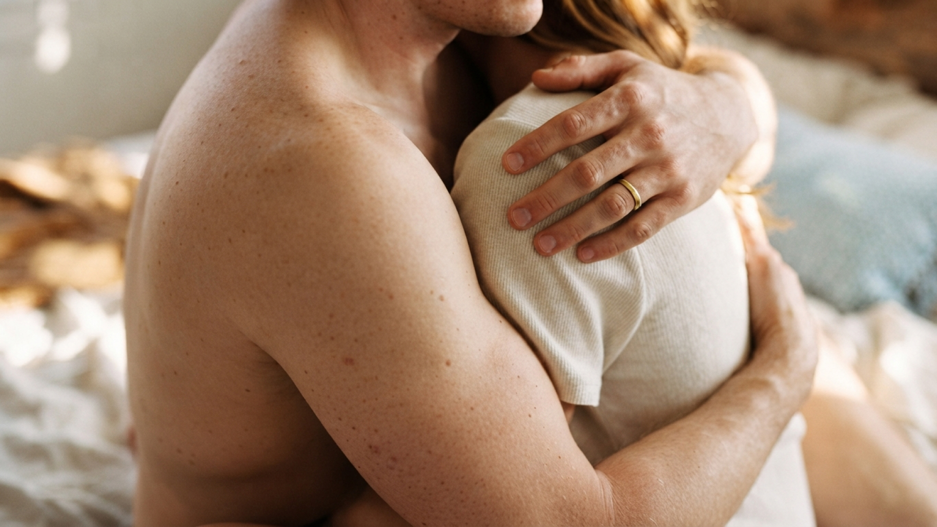 A shirtless man and a woman hug closely in bed, her hand on his shoulder showing a gold wedding ring