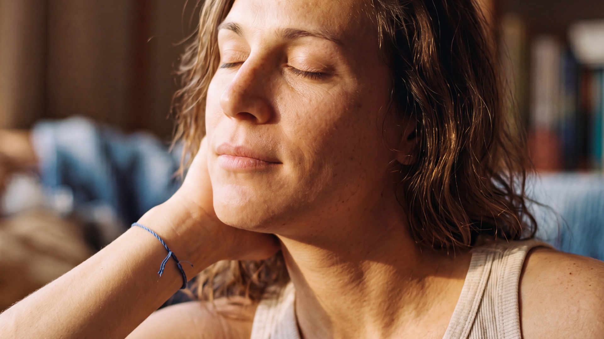 A woman with wavy brown hair rests her cheek on her hand with her eyes closed, calm and present in warm sunlight.