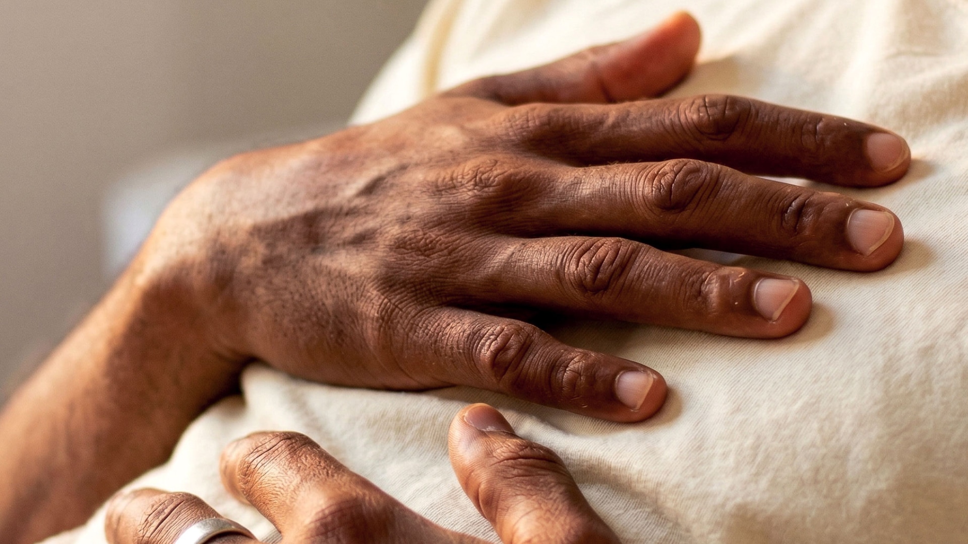 Close-up of a Black man resting his hand on his chest over a cream T-shirt