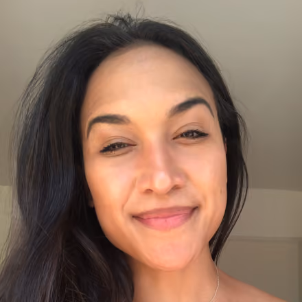 Close-up of a woman with long dark hair smiling gently in soft natural light against a plain light background.