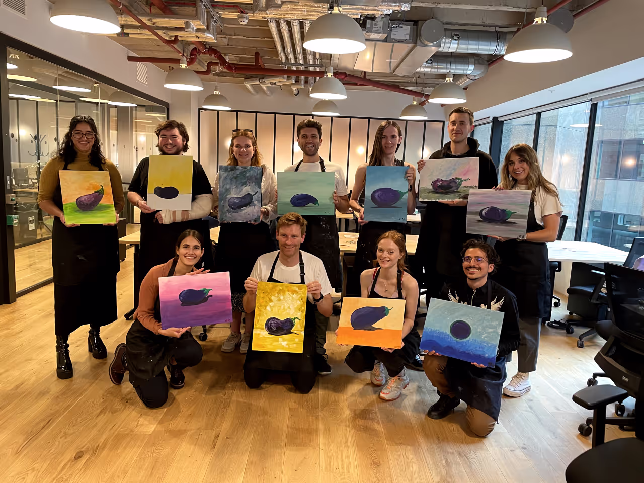 Group of eleven people in a bright room holding paintings of eggplants with different backgrounds, all dressed in black aprons.