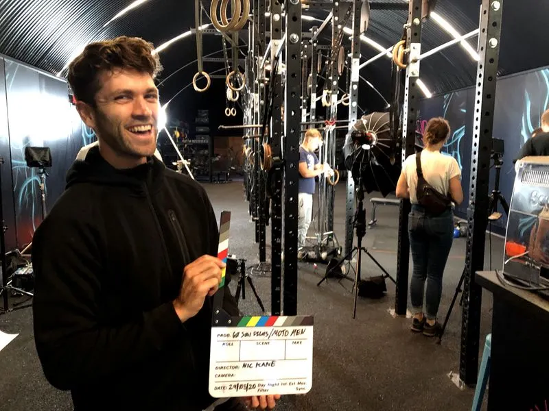 Smiling man in a black hoodie holding a film clapperboard inside a gym with workout equipment and crew members working in the background.