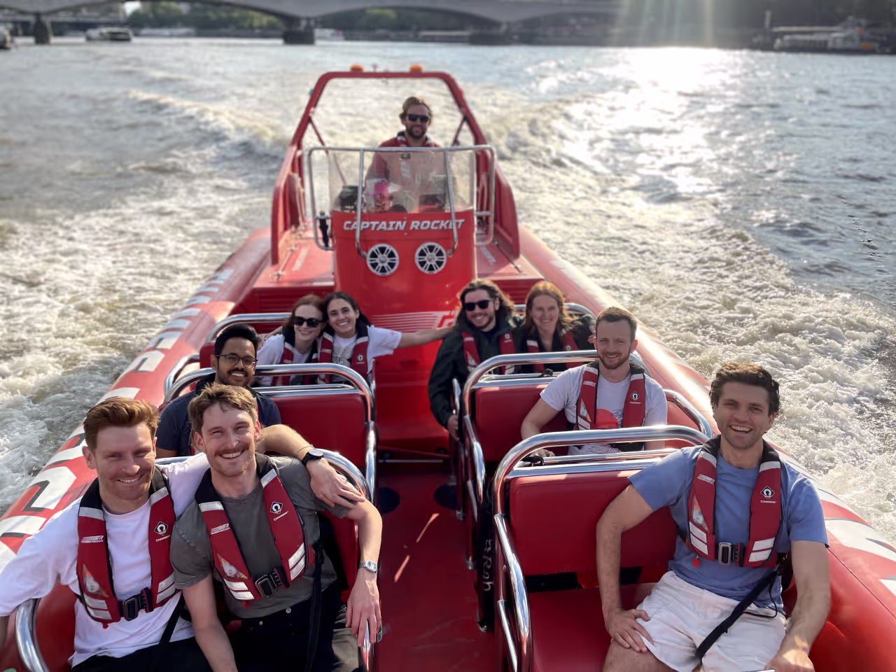 Group of nine people wearing life jackets smiling and sitting on a red Captain Rocket speedboat on a waterway with a bridge in the background.