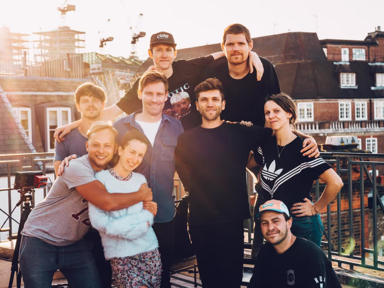 Group of nine young adults standing closely together on a rooftop with city buildings behind them, smiling and posing for the photo in casual clothing during daylight.
