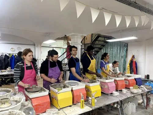 Six individuals standing side by side at potter's wheels in a studio, wearing aprons and shaping clay to make pottery with shelves and coats in the background.