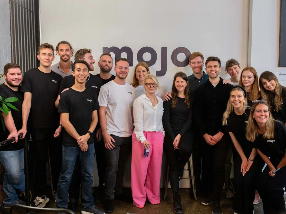 Group photo of smiling young adults standing indoors in front of a wall with the large text 'mojo'. Most wear casual black or white shirts with the mojo logo, with one person in a white top and bright pink pants holding a water bottle.