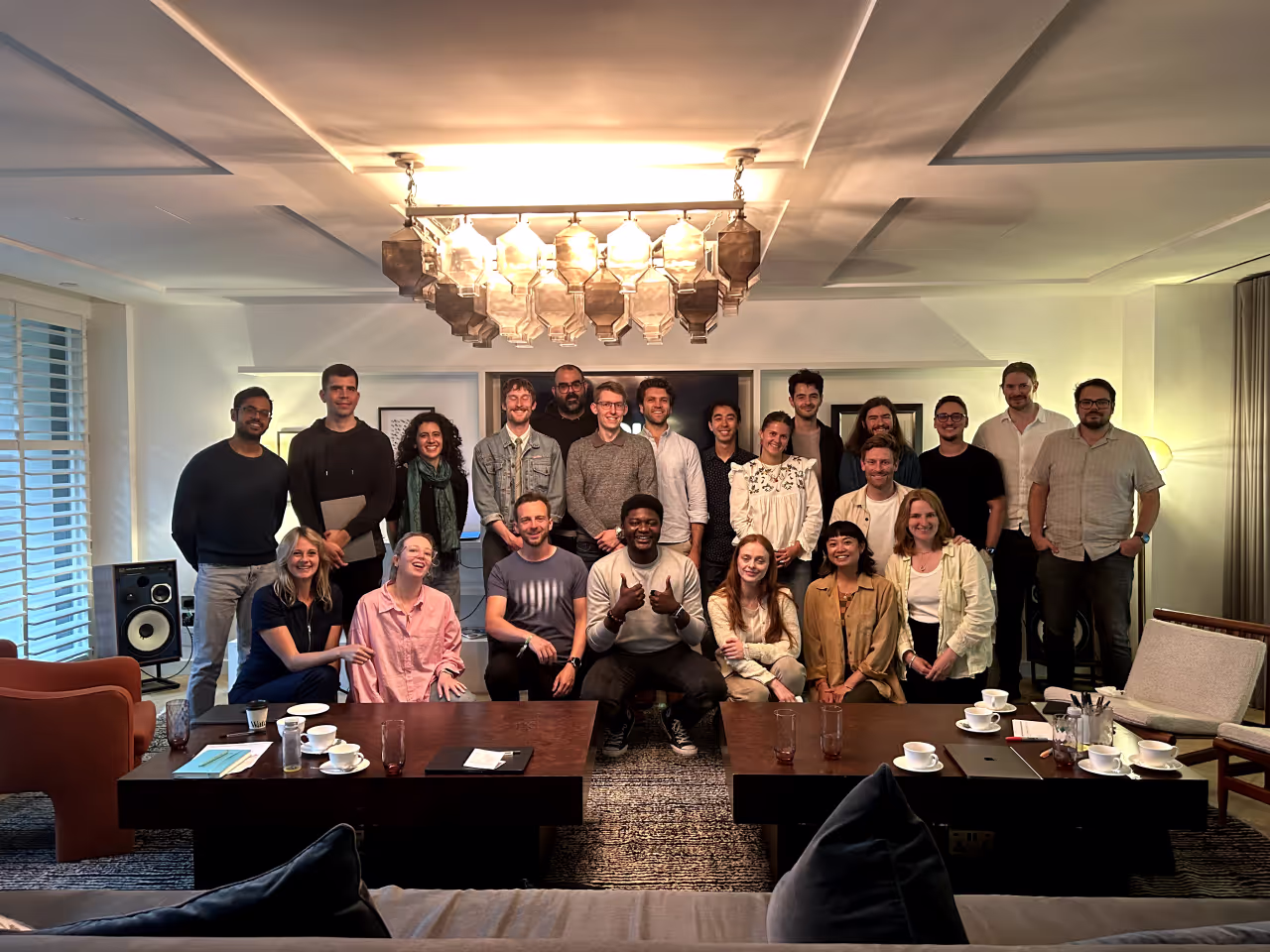 Group photo of 20 diverse people posing in a cozy, well-lit living room with modern decor, two coffee tables, cups, and a chandelier.