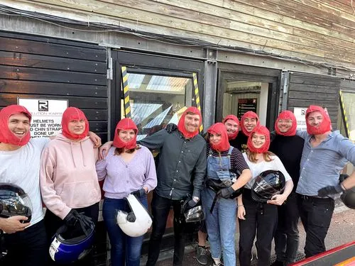 A group of ten people standing close together outside a building, all wearing red balaclava masks, some holding motorcycle helmets, smiling and posing for the photo.