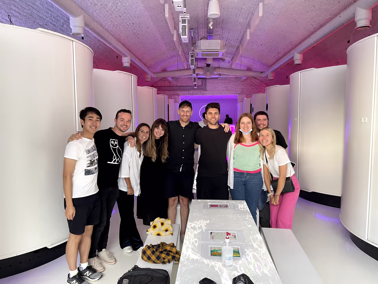 A group of nine diverse young adults smiling and posing together in a modern, softly lit indoor space with curved white walls and a long white table in front of them.
