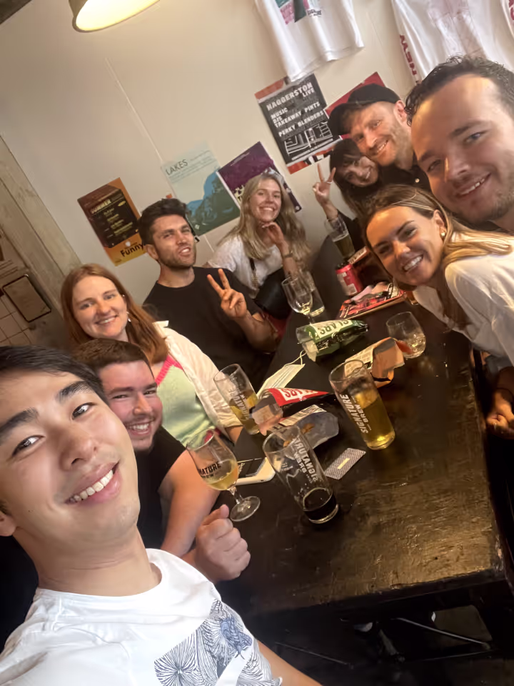 A group of eight young adults sitting around a table in a casual setting, smiling and posing for a selfie; several glasses and snack bags are on the table, and posters and t-shirts are visible on the wall behind them.
