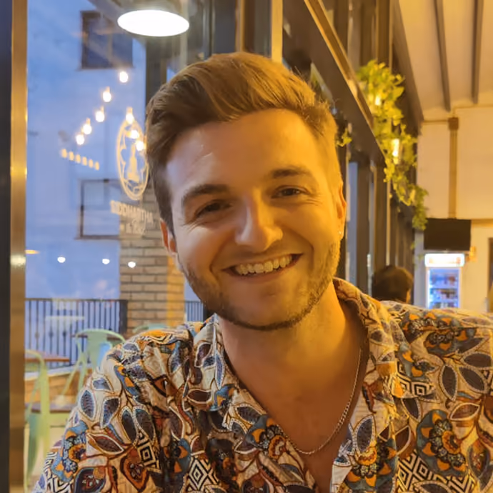 Smiling young man with short light brown hair and a trimmed beard, wearing a patterned shirt and a thin necklace, sitting indoors near glass windows with warm lighting and plants in the background.