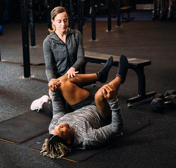 A woman in a gray hoodie assists a man lying on a mat in a gym, helping stretch his bent leg by holding his knee and foot.