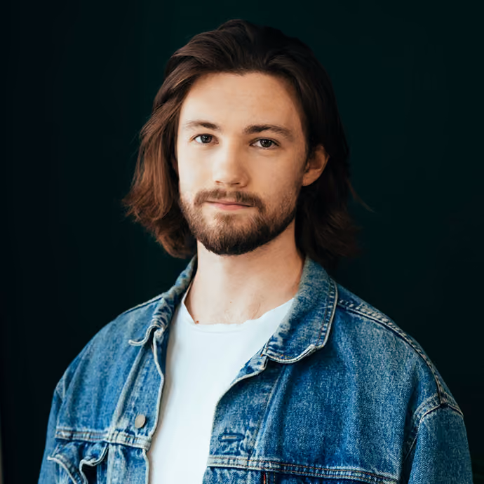 Young man with shoulder-length brown hair and a beard, wearing a white T-shirt and a blue denim jacket, against a dark background.
