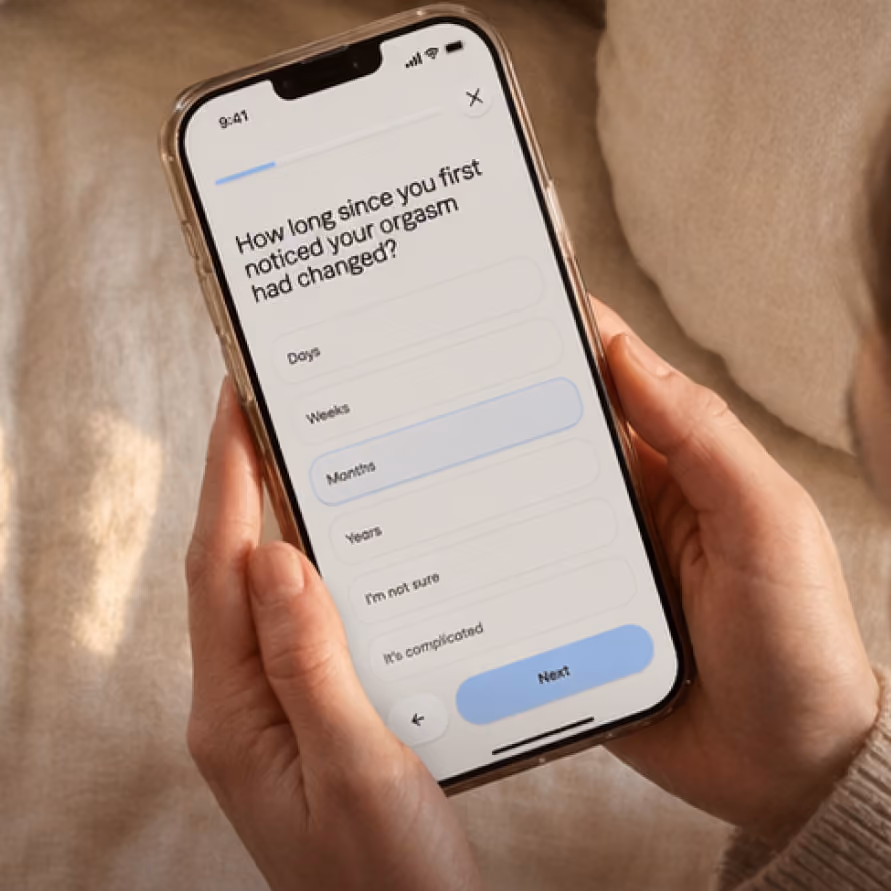 Smartphone screen displaying a health survey question asking, 'How long since you first noticed your orgasm had changed?' with answer options including Days, Weeks, Months (highlighted), Years, I'm not sure, and It's complicated, held by hands against a beige background.