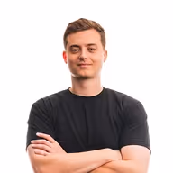 Young man with short light brown hair wearing a black t-shirt, standing with arms crossed and smiling slightly against a white background.