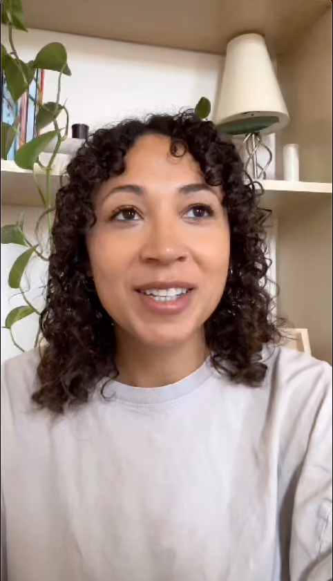 Smiling woman with curly hair wearing a light grey shirt sitting indoors with a plant and a lamp in the background.