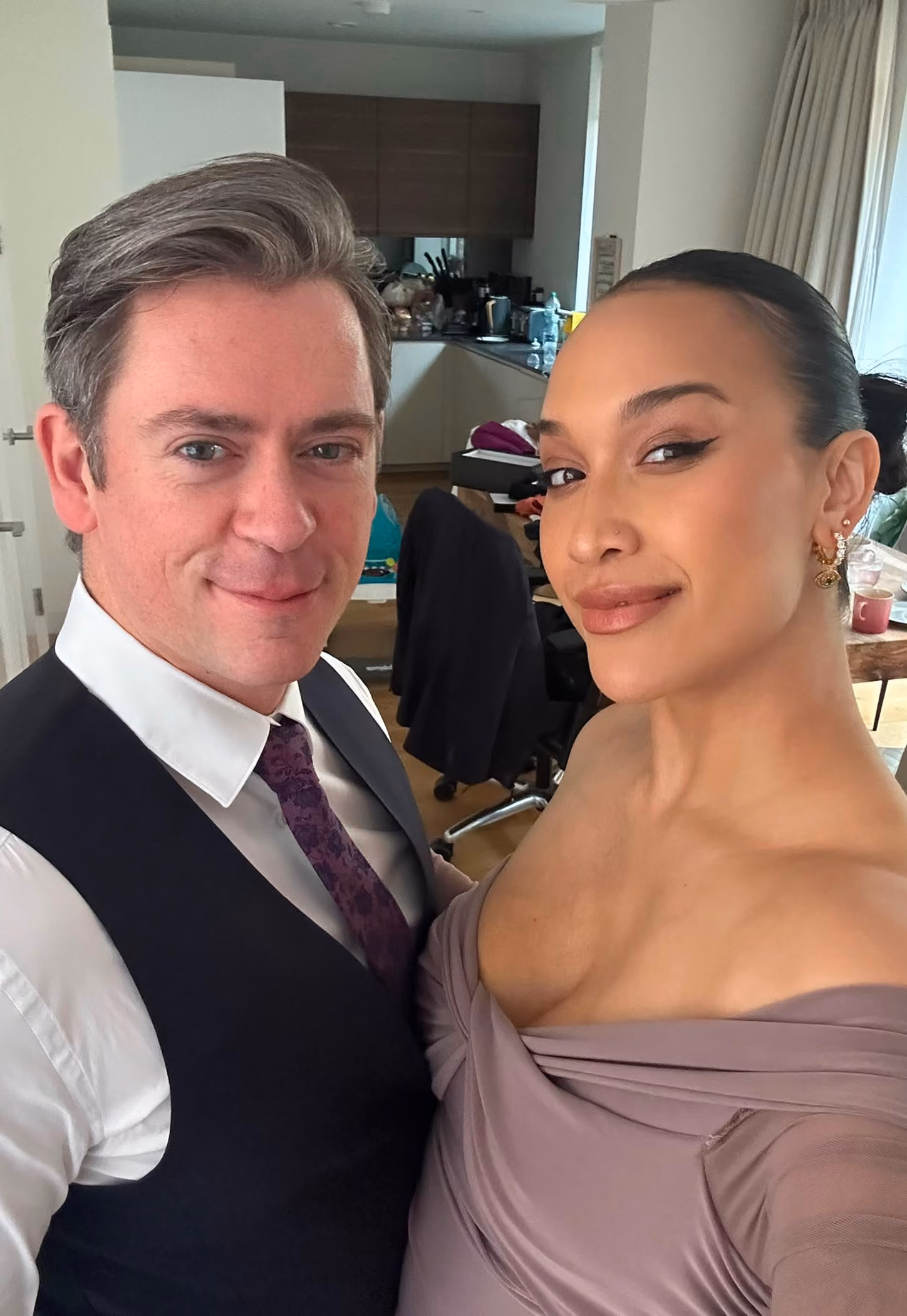 A man wearing a white shirt, purple tie, and dark vest stands next to a woman in a mauve off-shoulder dress with gold earrings, both smiling for a close-up selfie in a room with kitchen cabinets and a window with curtains.