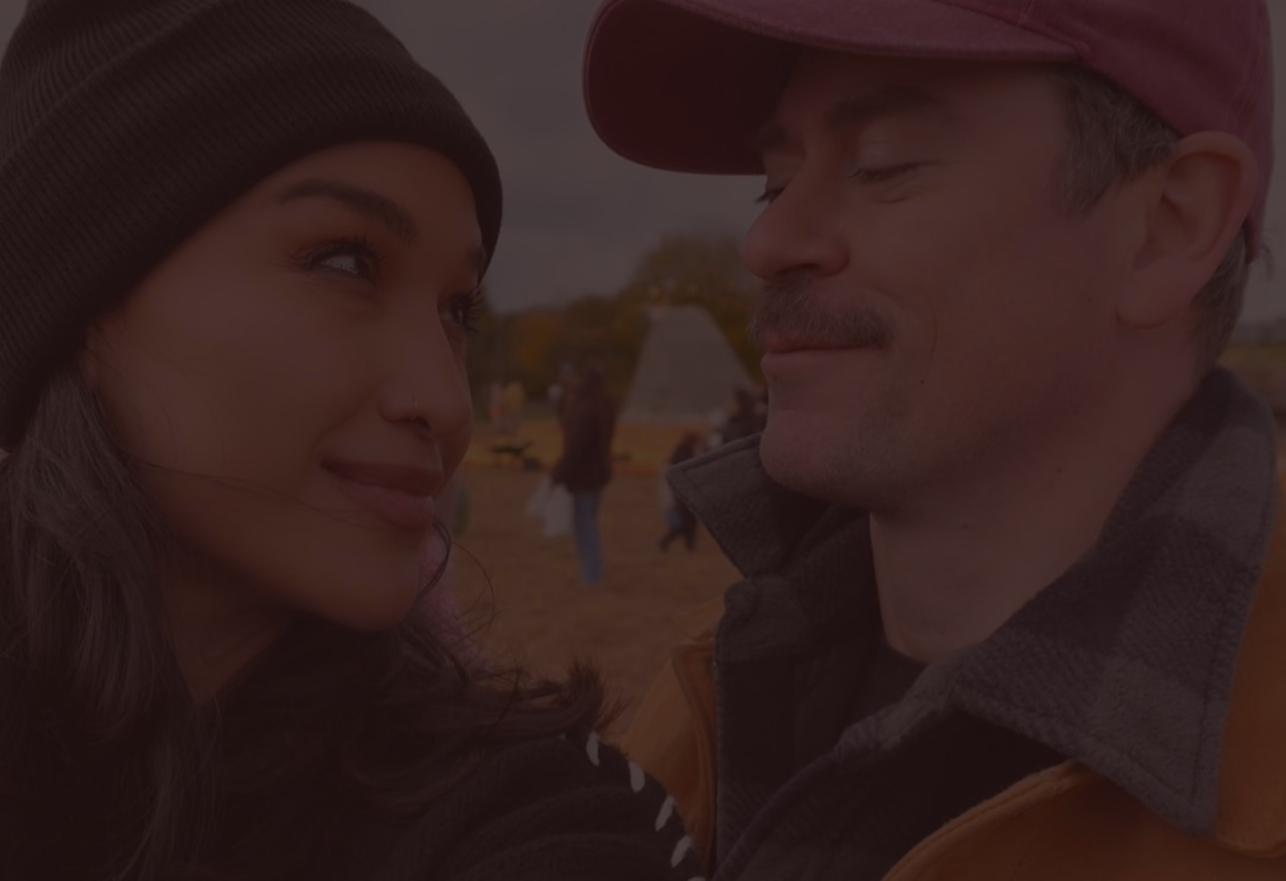 Close-up of a woman wearing a dark beanie looking and smiling at a man wearing a maroon cap and brown jacket with a checkered collar, both outdoors with blurred background.