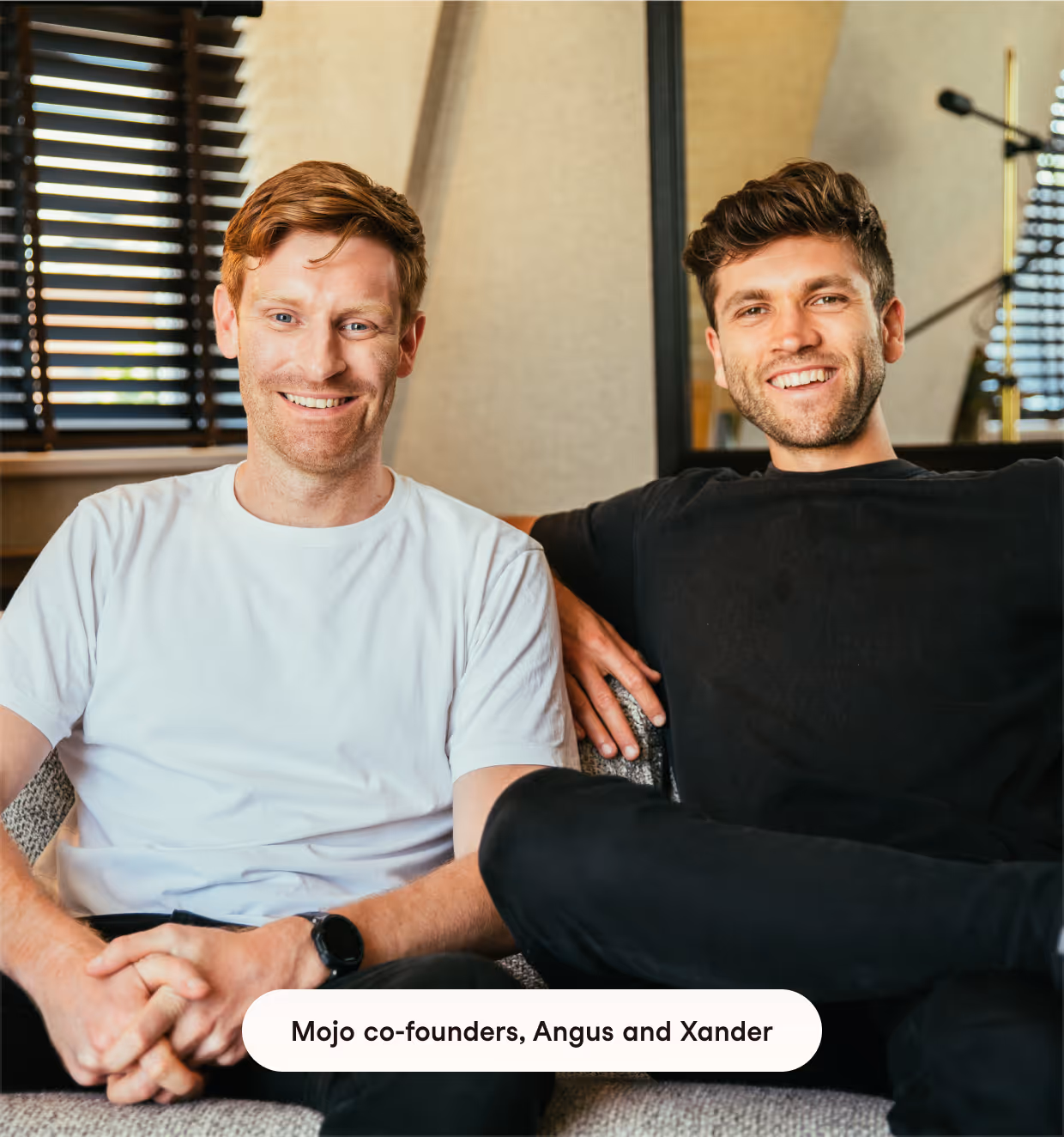 Two men sitting on a couch indoors, smiling at the camera; the man on the left has light hair and wears a white t-shirt, the man on the right has dark hair and wears a black shirt.