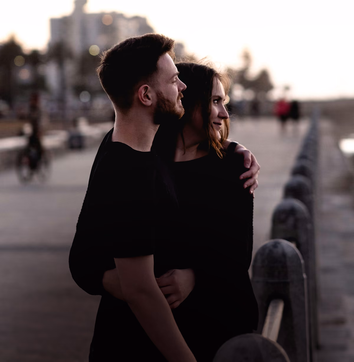 A young couple in black clothing embracing outdoors near a railing at dusk with blurred background of people and buildings.