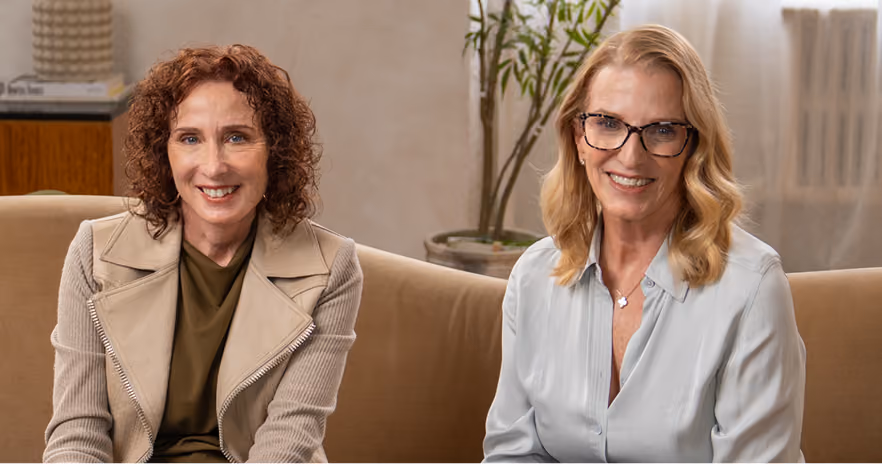 Two smiling women sitting on a beige sofa in a warmly lit room with a potted plant and window in the background.