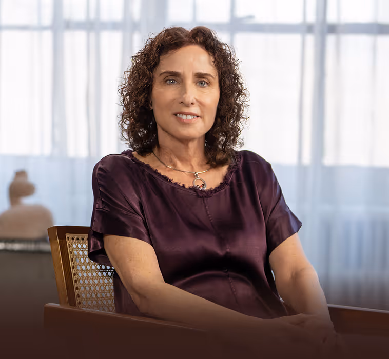 Woman with curly brown hair wearing a purple satin blouse and a silver necklace, seated on a wooden chair with woven backrest, smiling gently in a softly lit room with white curtains.