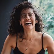 Young woman with curly dark hair laughing joyfully outdoors wearing a black tank top and a necklace, with blurred green foliage in the background.