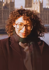 Young woman with curly hair and large glasses smiling outdoors in sunlight, wearing a dark coat and a black turtleneck sweater, with blurred buildings in the background.