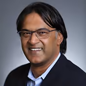 Portrait of a middle-aged man with medium skin tone, black hair, glasses, and a dark blazer smiling against a gray background.