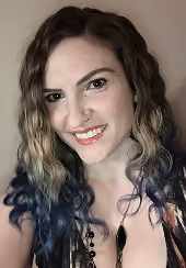 Smiling woman with wavy hair dyed dark at the ends, wearing a nose ring and a black necklace against a plain beige background.