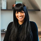 Smiling woman with long straight black hair and bangs, wearing a black top, seated in a room with a blurred background.