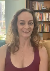 Smiling woman with curly hair wearing a maroon sleeveless top sitting on a couch in front of a bookshelf filled with books.