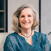 Smiling middle-aged woman with gray hair wearing a teal patterned blouse and a necklace, seated indoors in a softly lit room.