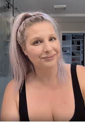 Woman with light pink hair tied in a high ponytail, wearing a black tank top, smiling softly in an indoor setting with shelves in the background.