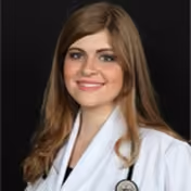Smiling young woman with long brown hair wearing a white medical coat and a stethoscope around her neck against a black background.