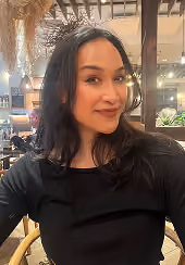 Woman with medium-length dark hair wearing a black top, sitting indoors with warm lighting and plants in the background.