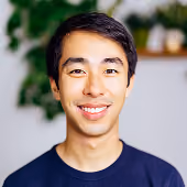 Smiling young Asian man with short dark hair wearing a dark blue shirt with a blurred green plant and wooden shelf in the background.
