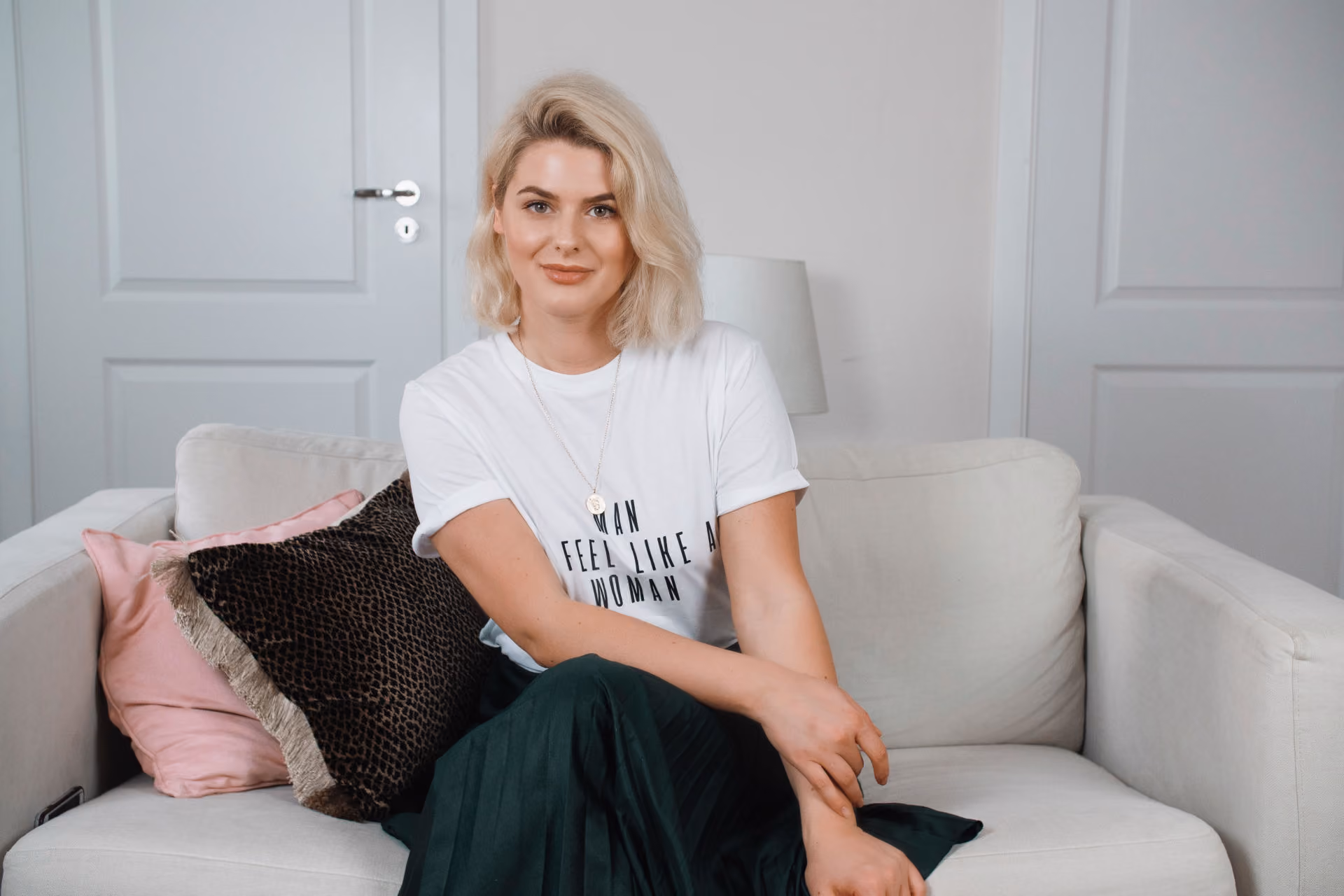 A blonde woman with shoulder-length hair sitting on a light-colored sofa in a modern room, wearing a white T-shirt and dark skirt, smiling gently with crossed legs and arms resting on her lap, with decorative pillows behind her on the sofa and closed white doors in the background.