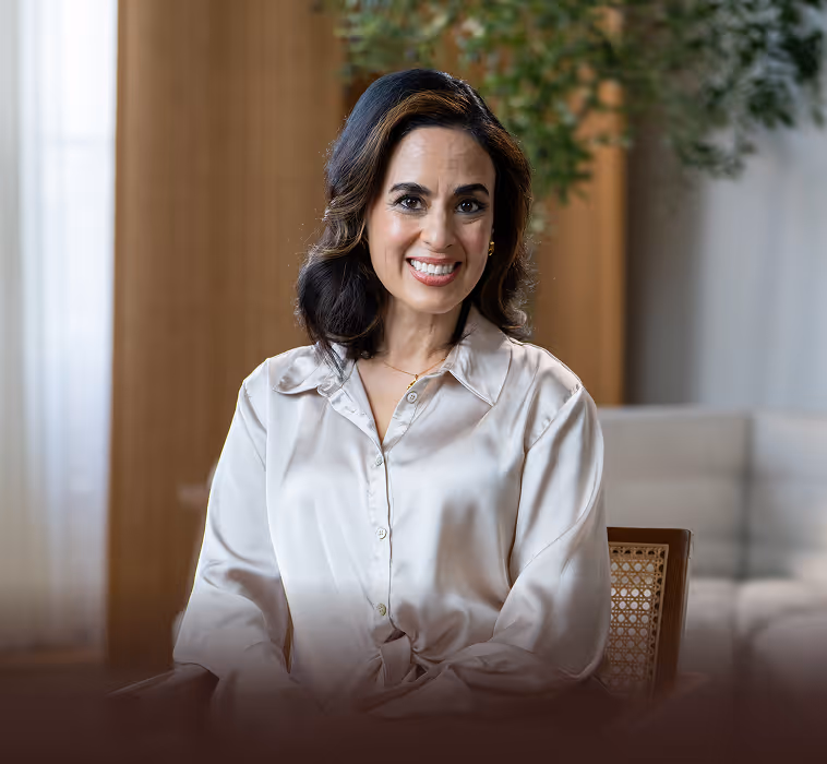 Smiling woman with dark hair wearing a cream-colored satin blouse, seated indoors with blurred plants and furniture in the background.